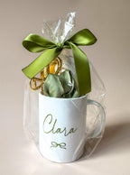 Coffret cadeau mug personnalisé femme avec nœud vert sauge et accessoires cheveux. Mug en céramique blanche personnalisé avec prénom, présenté avec ruban satiné vert sauge. Coffret bien-être femme avec mug personnalisé, chouchou en satin et pinces dorées.