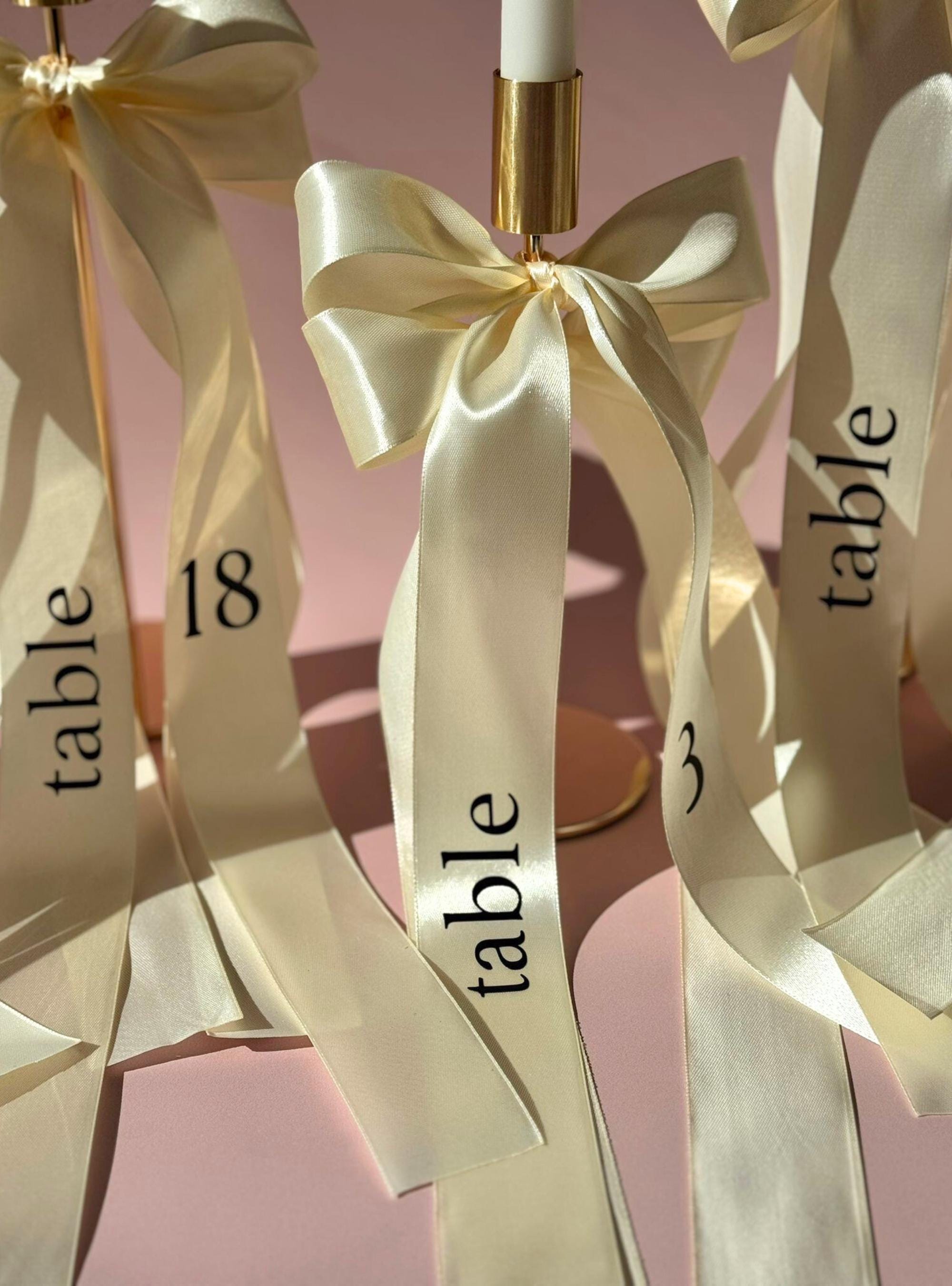 Wedding Table Number Display Ribbon Centerpiece Design Cream Satin Material Elegant Bow Detail Gold Hardware Accent Reception Seating Marker Formal Event Decoration Luxury Wedding Accessory Customizable Font Option Table Identification
