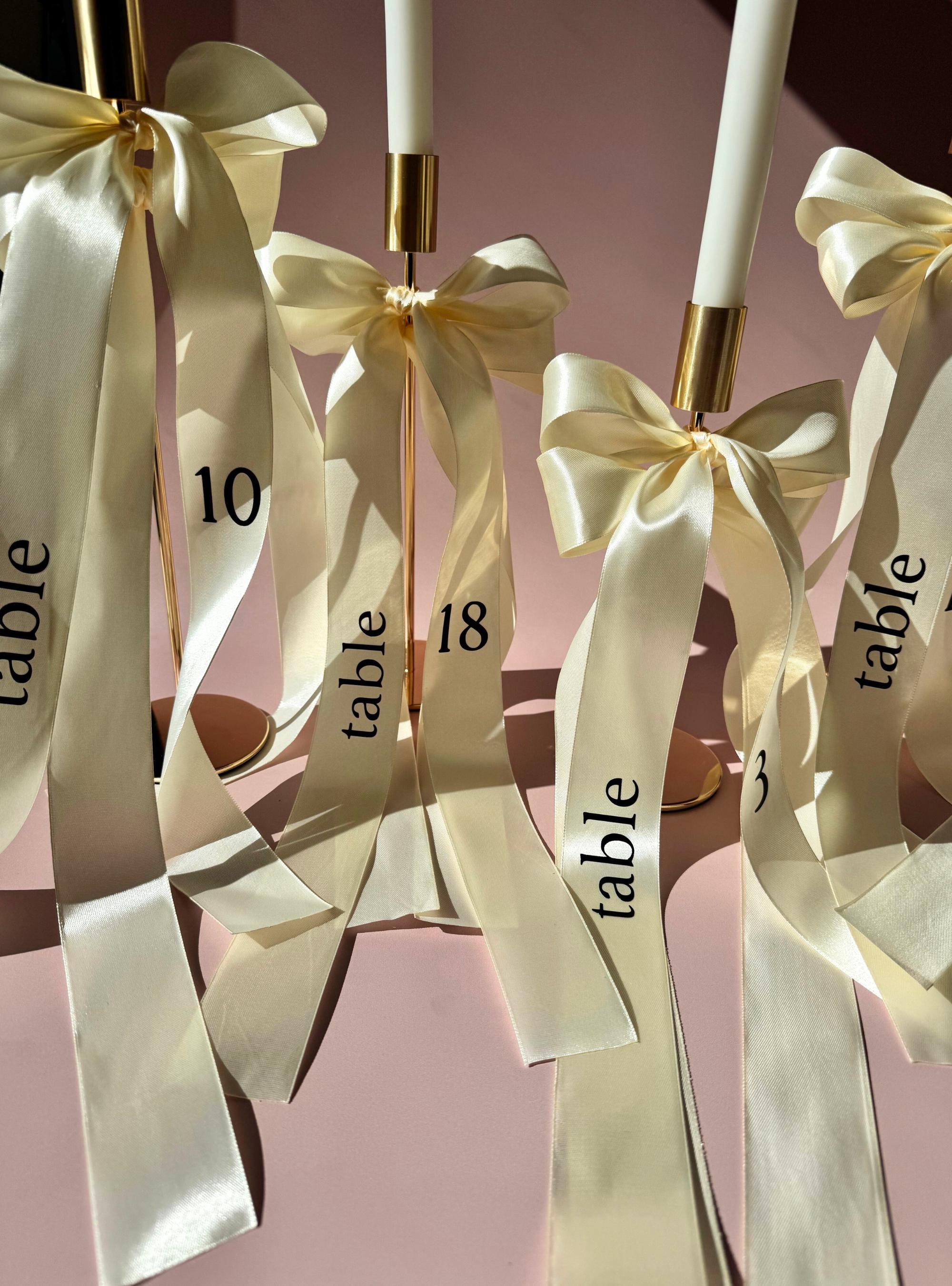 Elegant Centerpiece Ribbon Wedding Table Number Display Satin Bow Design Reception Decoration Cream Color Material Gold Hardware Accent Formal Event Marker Beautiful Seating Arrangement Customizable Text Option Luxury Wedding Accessory